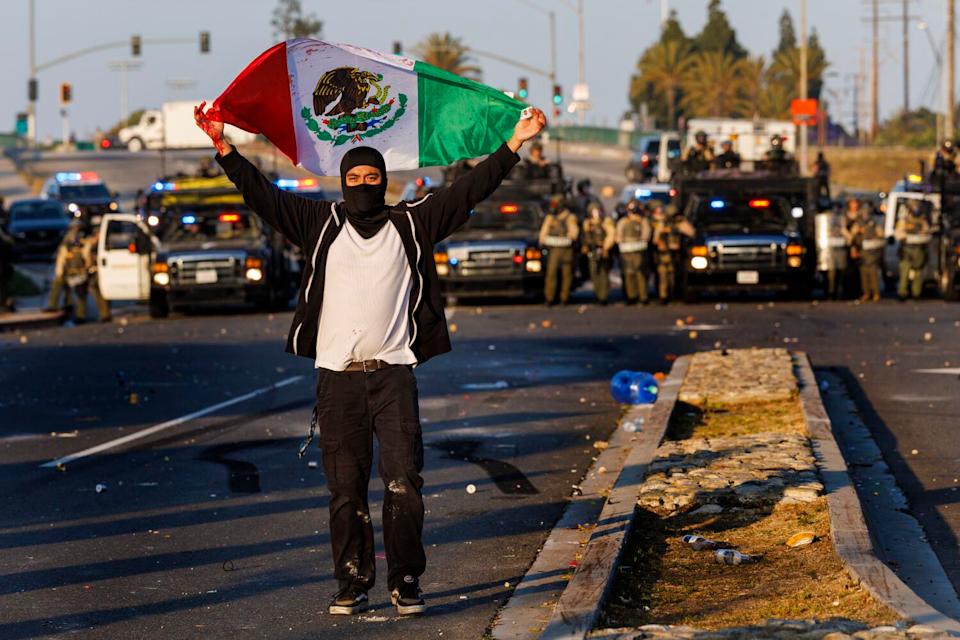 A protester carries a Mexican flag as L.A. County sheriff's deputies form a law enforcement line far behind him.