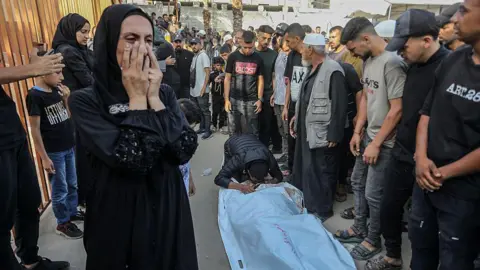 Getty Images A body bag lies on a ground while a man kneels to kiss it. Others gather round while a woman covers her face and cries. 