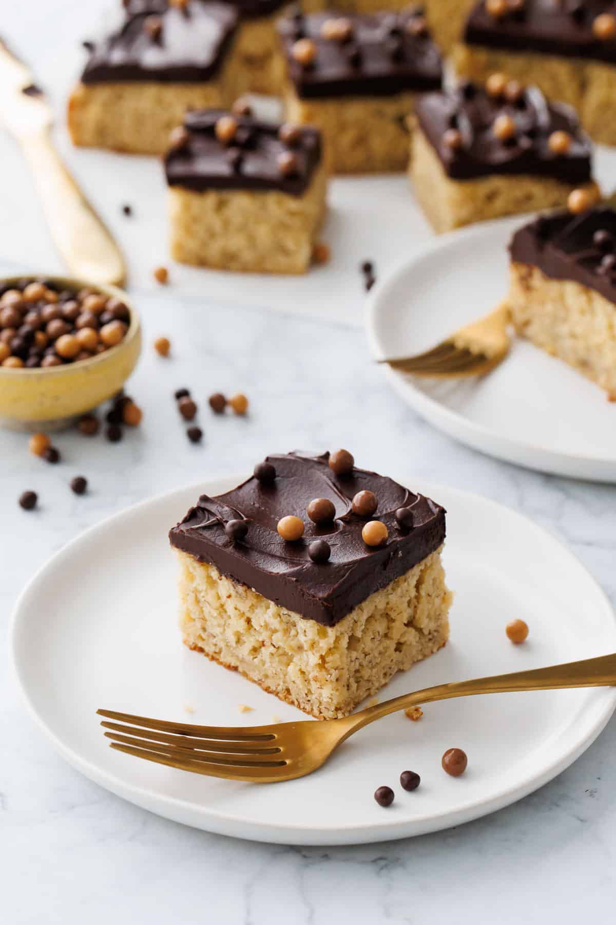 Square of cut Brown Butter Banana Cake with Chocolate Fudge Frosting on a white plate with gold fork, more slices in the background.