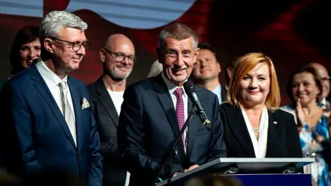 Anadolu via Getty Images Andrej Babis holds a press conference at ANO headquarters on 4 October 2025, wearing a blue suit and red tie. He is surrounded by people including a man in a suit and a woman in a suit