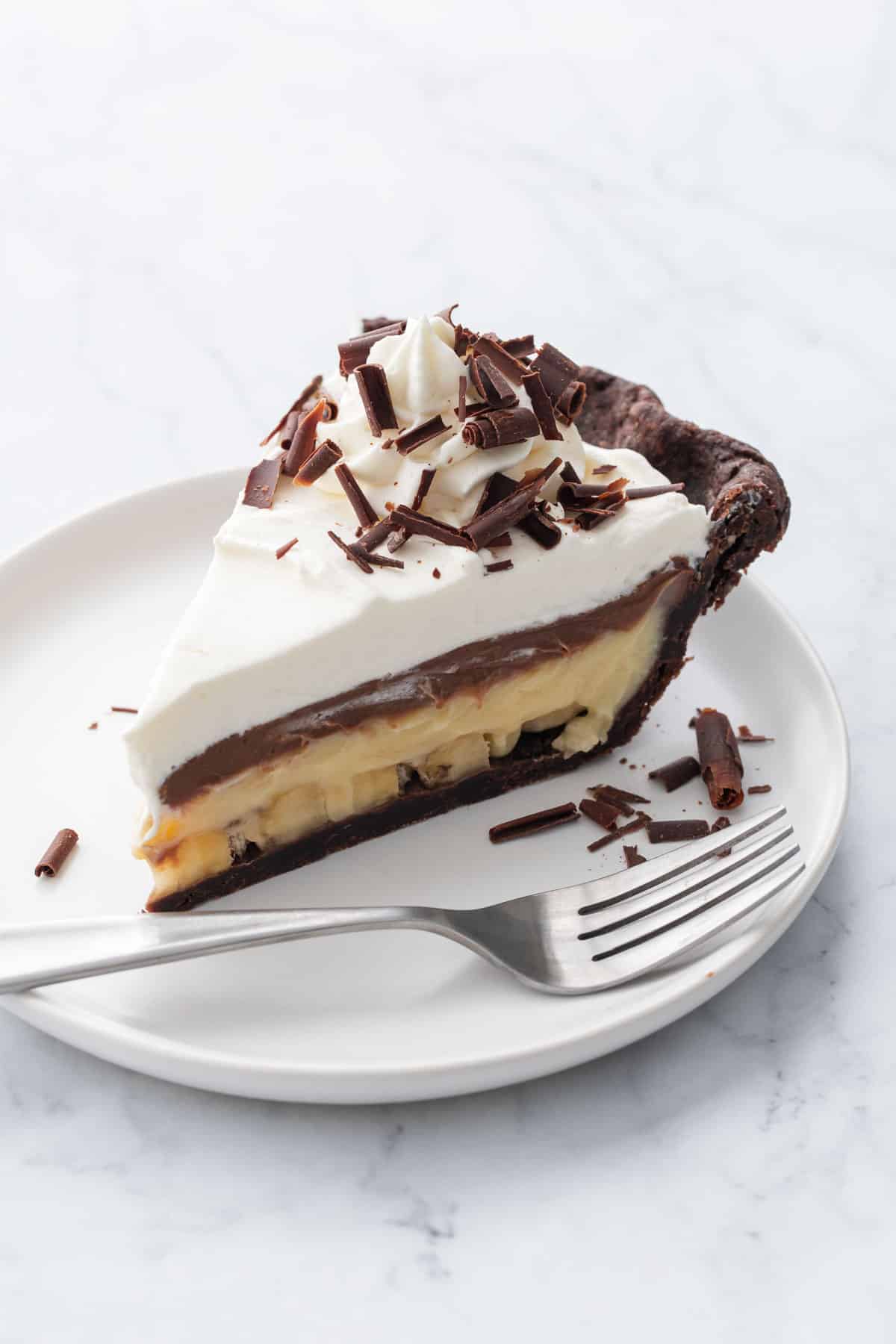Slice of chocolate banana cream pie on a white plate on a marble background with silver fork.