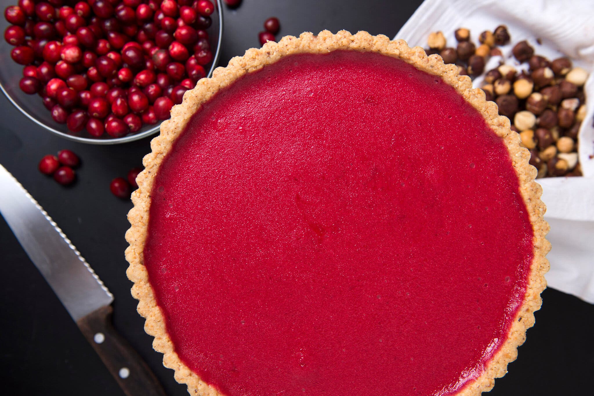 An overhead view of a bright red tart. To the side are a bowl of cranberries, a pile of nuts on a white cloth and a knife.