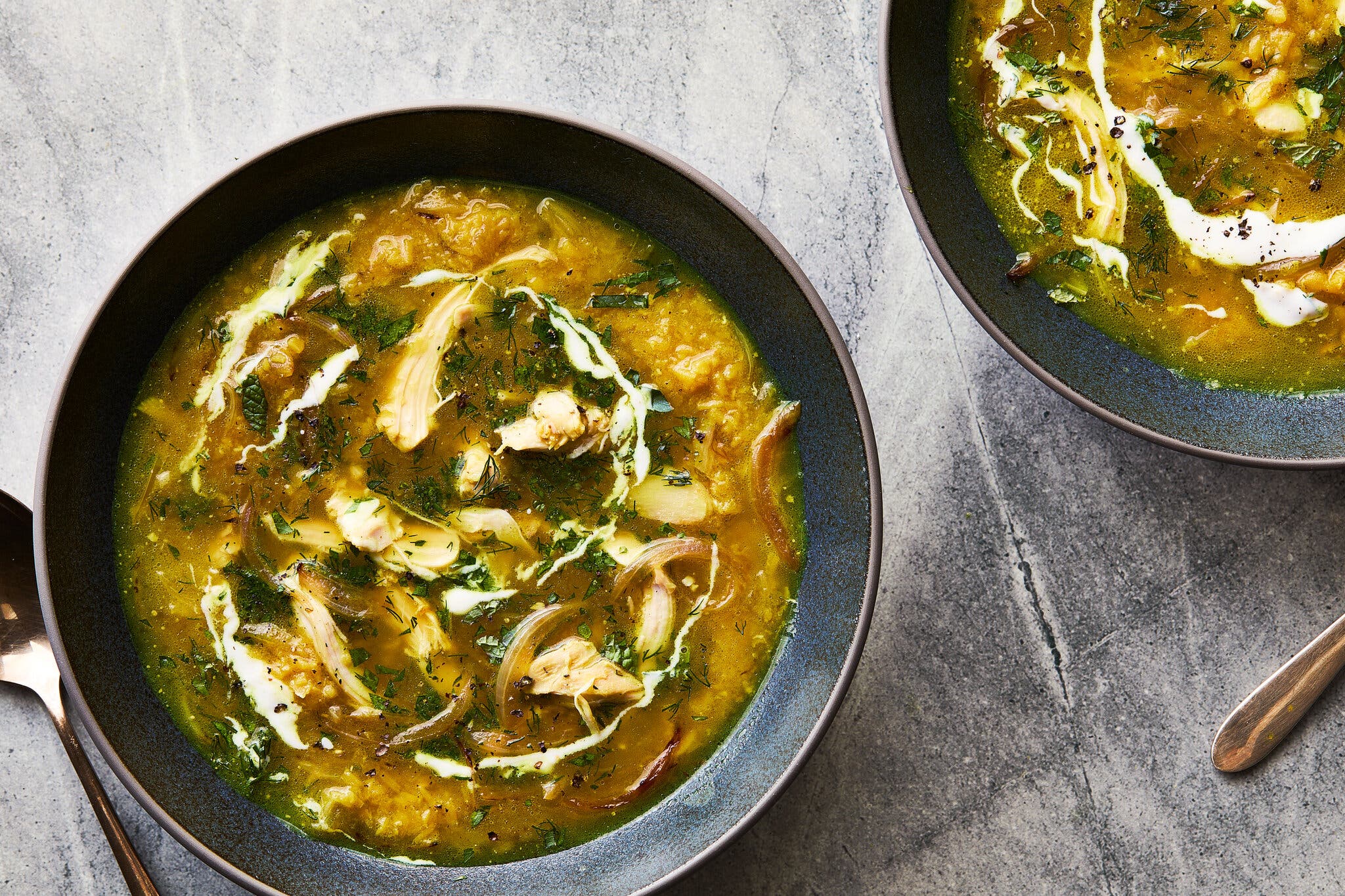 A dark blue bowl filled with a rich yellow chicken soup, topped with shredded chicken, a white yogurt swirl and fresh herbs.