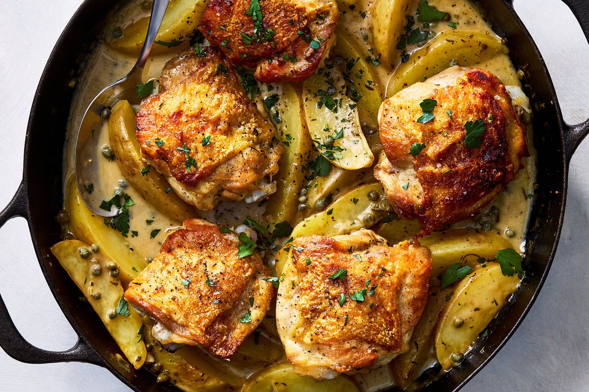An overhead image of chicken thighs atop sauced potato wedges in a cast-iron skillet.