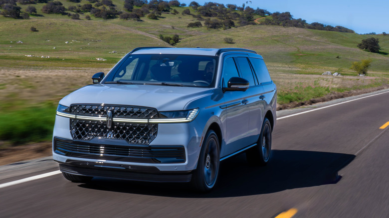 A front three quarters shot of a light blue Navigator driving on a two-lane road in front of rolling green hills with shrubs
